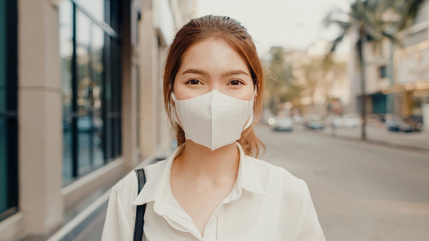 A person, wearing a white face mask and shirt, stands on an urban street lined with buildings and palm trees. The background is slightly blurred, capturing the serene yet bustling atmosphere—a subtle nod to avoiding that mouth breather vibe in San Antonio, TX