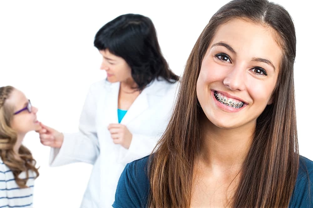 Fiesta Orthodontics A teen girl with braces, often curious about the cost, smiles brightly in the foreground. In the background, a doctor attentively examines a young child wearing glasses in San Antonio, TX