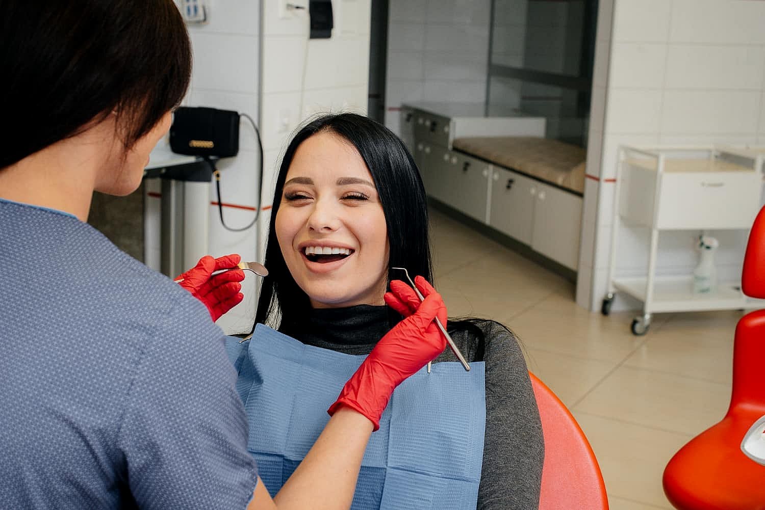 A dentist wearing red gloves prepares to examine a smiling patient's teeth, checking for signs of tongue thrust in the cozy dental office. The patient sits comfortably in a dental chair, sporting a blue bib in San Antonio, TX