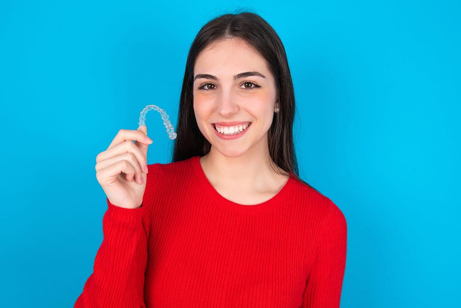 A person in a red shirt holds up a clear Invisalign dental aligner after understanding Invisalign cost, smiling against a blue background in San Antonio, TX