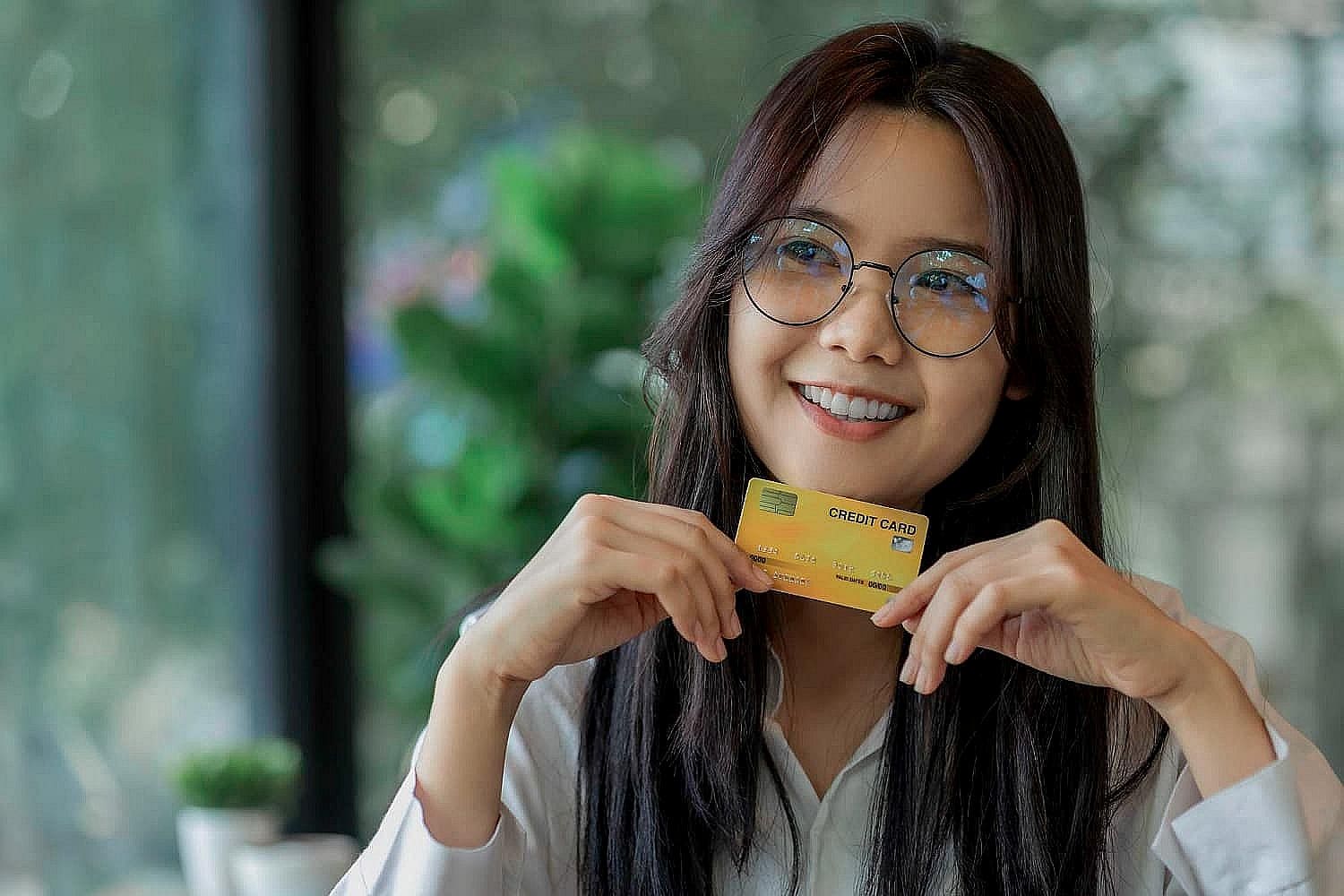 Woman with glasses smiling, holding a yellow credit card, seated indoors with blurred greenery in the background, planning her next purchase with the help of a flexible braces payment plan in San Antonio, TX
