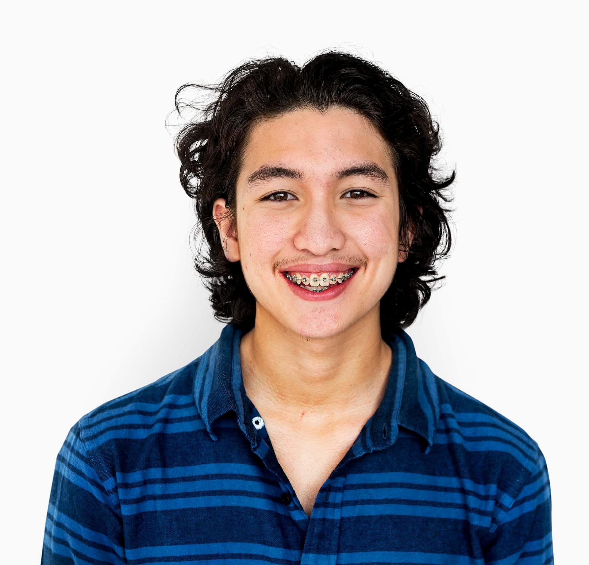 An adult with long hair and braces, donning a blue striped shirt, smiles warmly against a plain white background in San Antonio, TX