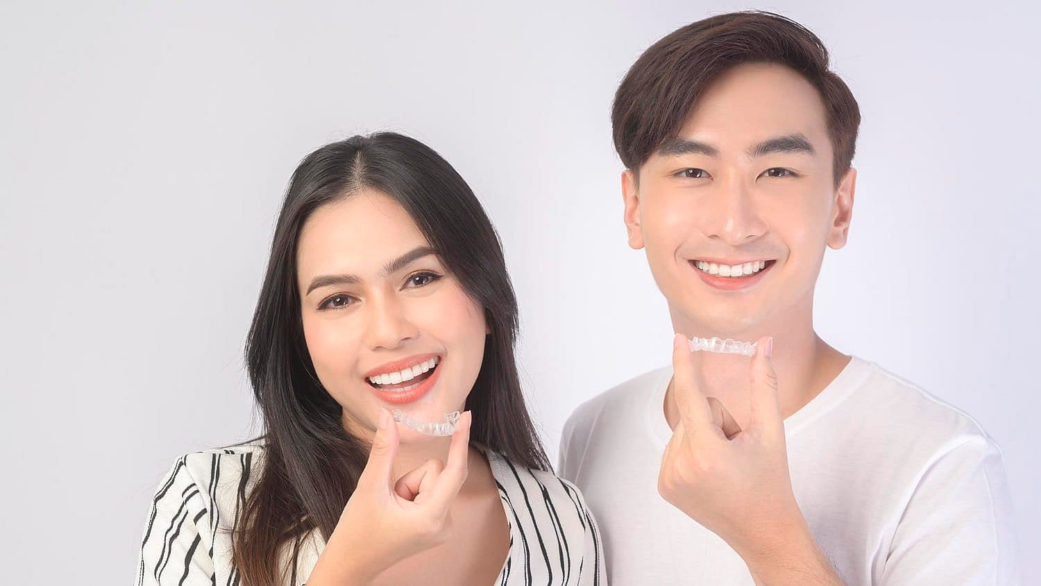 A man and a woman smile while holding their Invisalign clear dental aligners. The woman wears a striped top, and the man sports a white T-shirt in San Antonio, TX