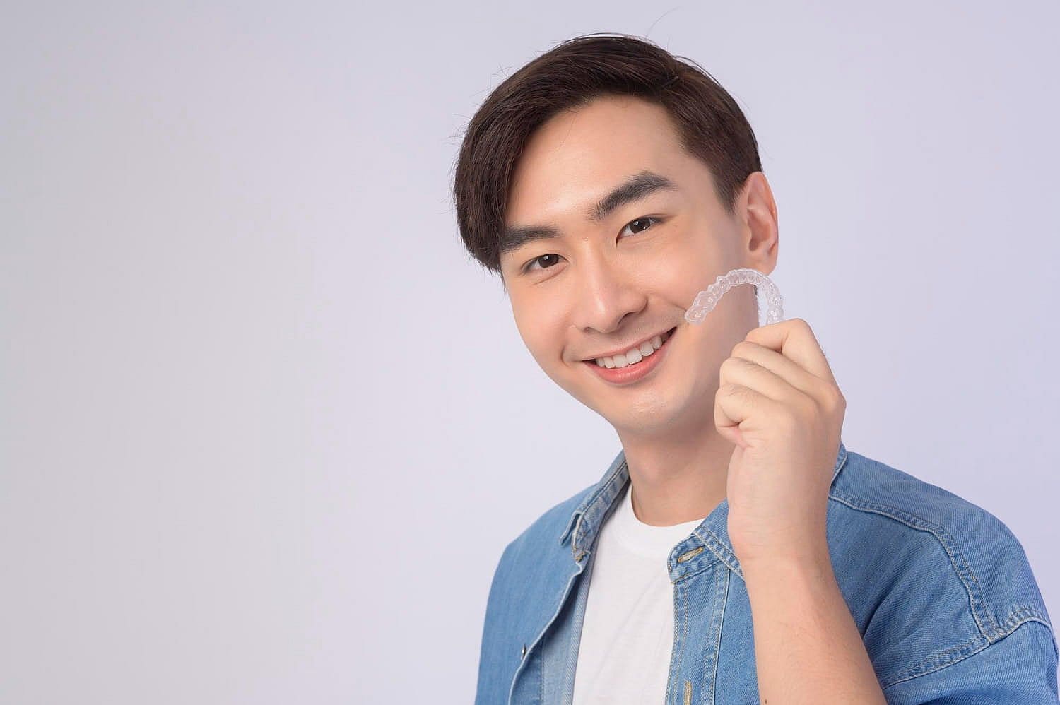 Person smiling and holding a clear dental aligner, wondering if Invisalign might be covered by insurance. They're dressed casually in a blue shirt layered over a white t-shirt in San Antonio, TX