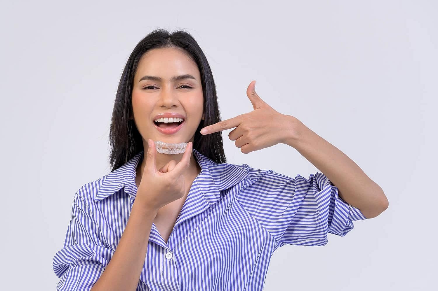 A person in a striped shirt smiles while holding a clear dental aligner, pointing proudly at it. They're excited about their Invisalign treatment, made even easier with an affordable payment plan in San Antonio, TX