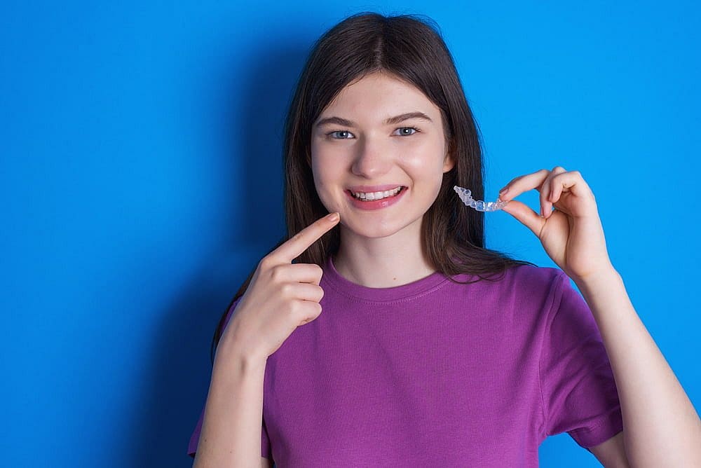 Young woman in a purple shirt holding a clear dental aligner, smiling, and pointing to her teeth against a blue background, highlighting the benefits of Invisalign vs braces in San Antonio, TX