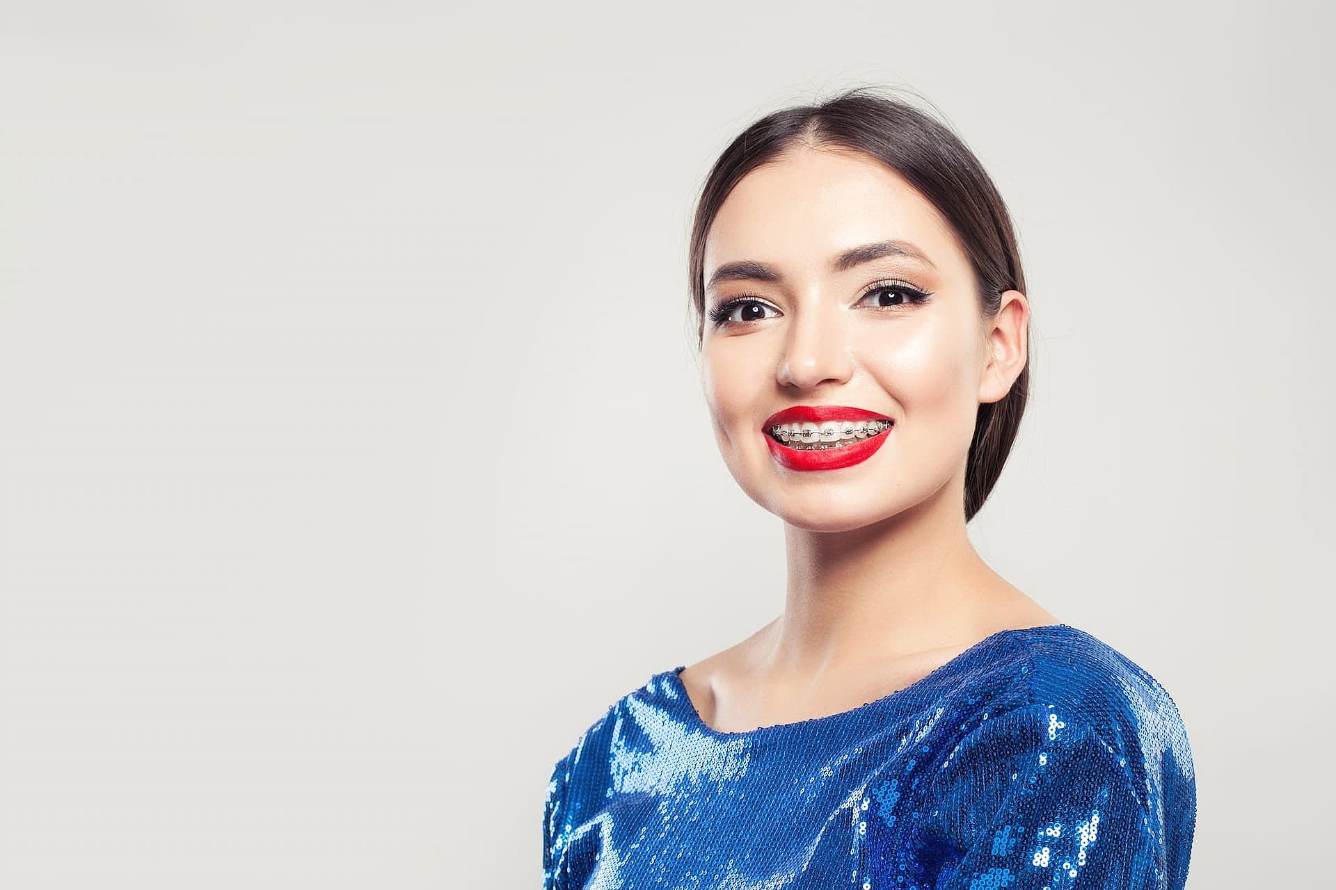 Woman with braces smiling confidently, showcasing the benefits of adult orthodontics. She is wearing a blue sequined top against a plain background in San Antonio, TX