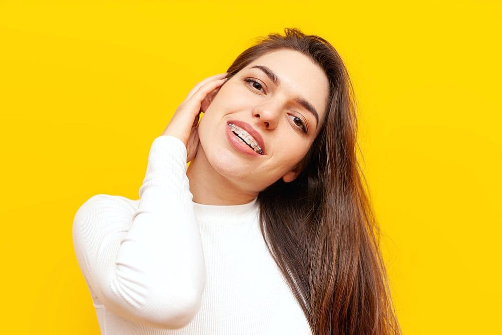 A person with long hair and clear braces smiles against a bright yellow background, wearing a white top in San Antonio, TX