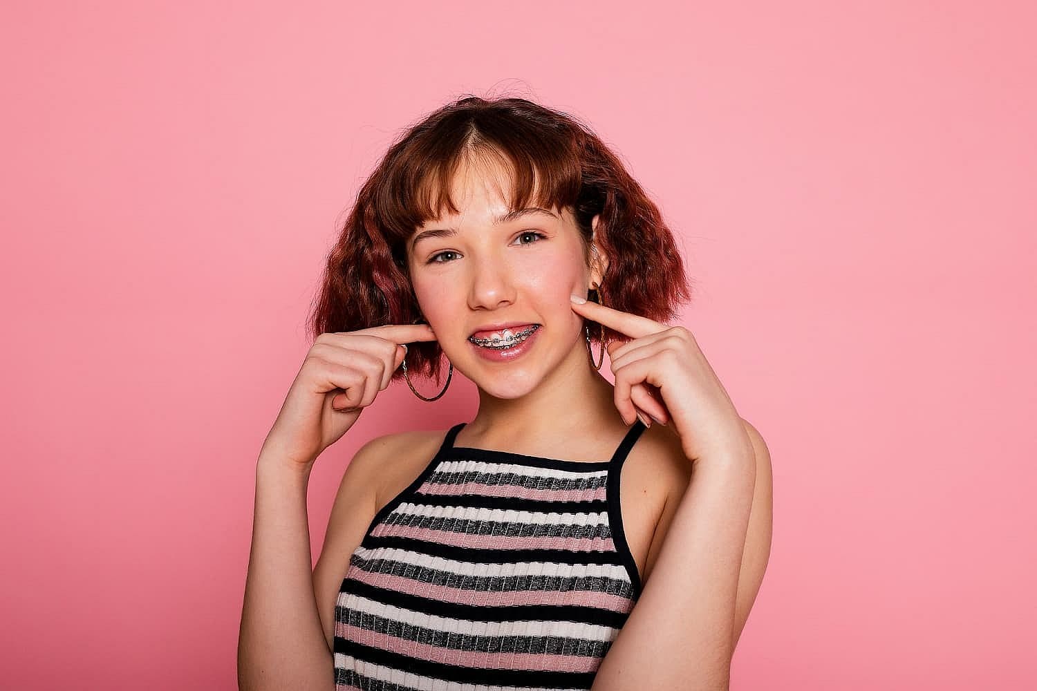 A person with braces smiles and points to their cheeks, proudly embracing the journey of Invisalign vs Braces. They have short brown hair and are wearing a striped sleeveless top against a pink background in San Antonio, TX
