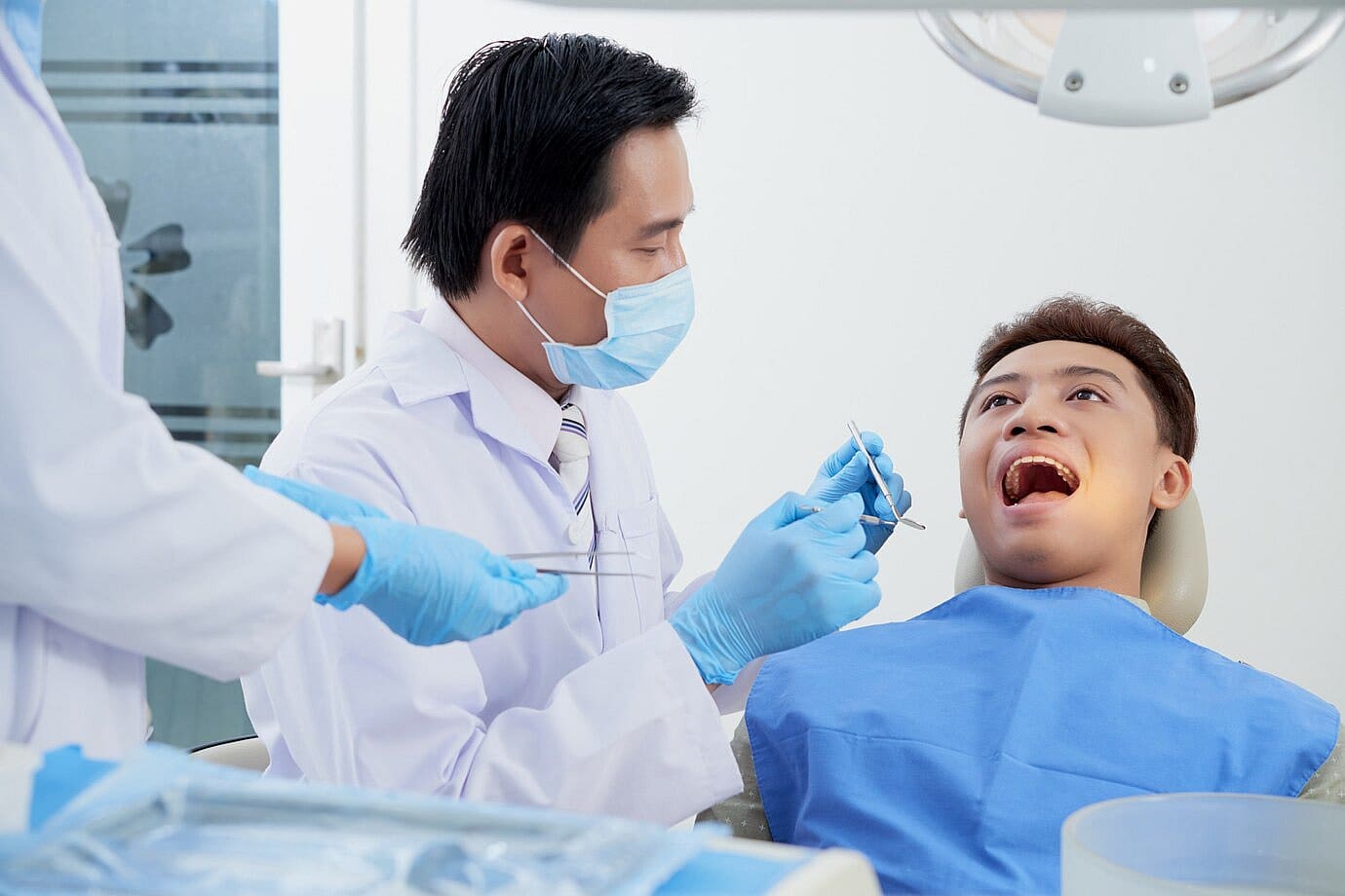 A dentist in a mask examines a patient's open mouth, checking for tongue thrust issues with dental tools, assisted by another person, in a clinical setting in San Antonio, TX