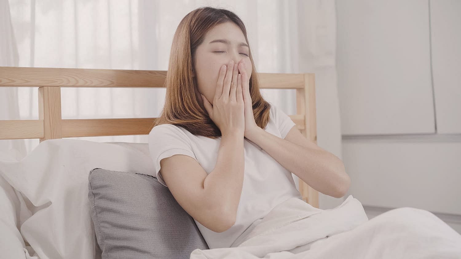 A person sitting up in bed yawning, hands gently covering their mouth, surrounded by white bedding and light-colored pillows—an image that could depict a serene moment for any quintessential mouth breather in San Antonio, TX