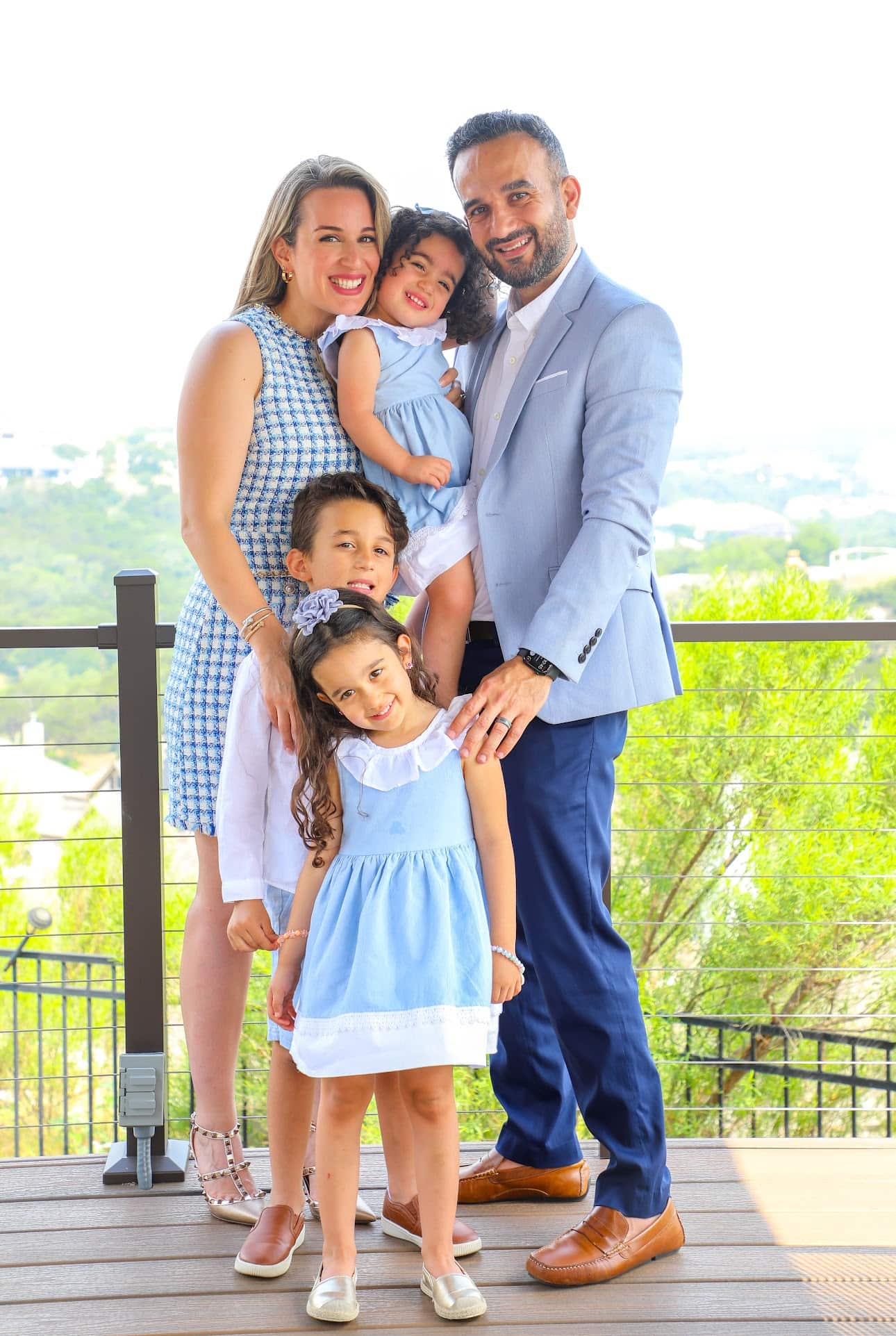 A family of five, with Dr. Fateh Arslan and Dr. Ayla Idilbi—orthodontics experts from Fiesta Orthodontics—poses outdoors. The parents stand behind their three children, all dressed in blue and white outfits. Trees and a serene landscape provide a picturesque backdrop for this cherished moment in San Antonio, TX.