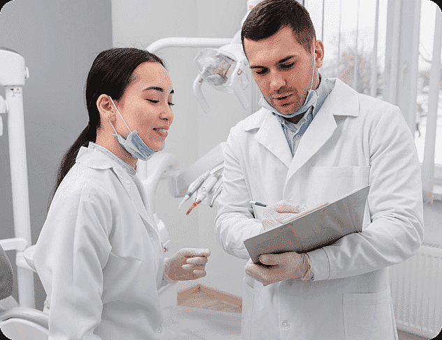 Two dentists in white coats and masks discuss a patient chart in a dental office, planning the best approach for adult orthodontics amidst the backdrop of advanced dental equipment in San Antonio, TX