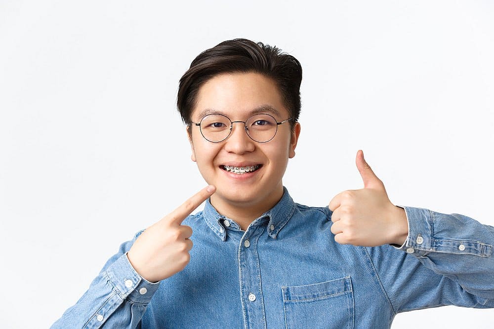 An adult sporting glasses and braces smiles confidently, pointing to their gleaming adult braces and giving a thumbs-up. Dressed in a denim shirt, they stand out against the plain background with effortless charm in San Antonio, TX