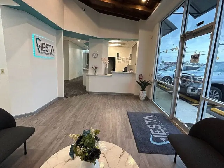 The lobby at Fiesta Orthodontics in East San Antonio, TX features a reception desk, indoor plants, a wall sign displaying the practice’s name, comfortable seating, and large windows overlooking the parking lot. The lobby at Fiesta Orthodontics in East San Antonio, TX features a reception desk, indoor plants, a wall sign displaying the practice’s name, comfortable seating, and large windows overlooking the parking lot.