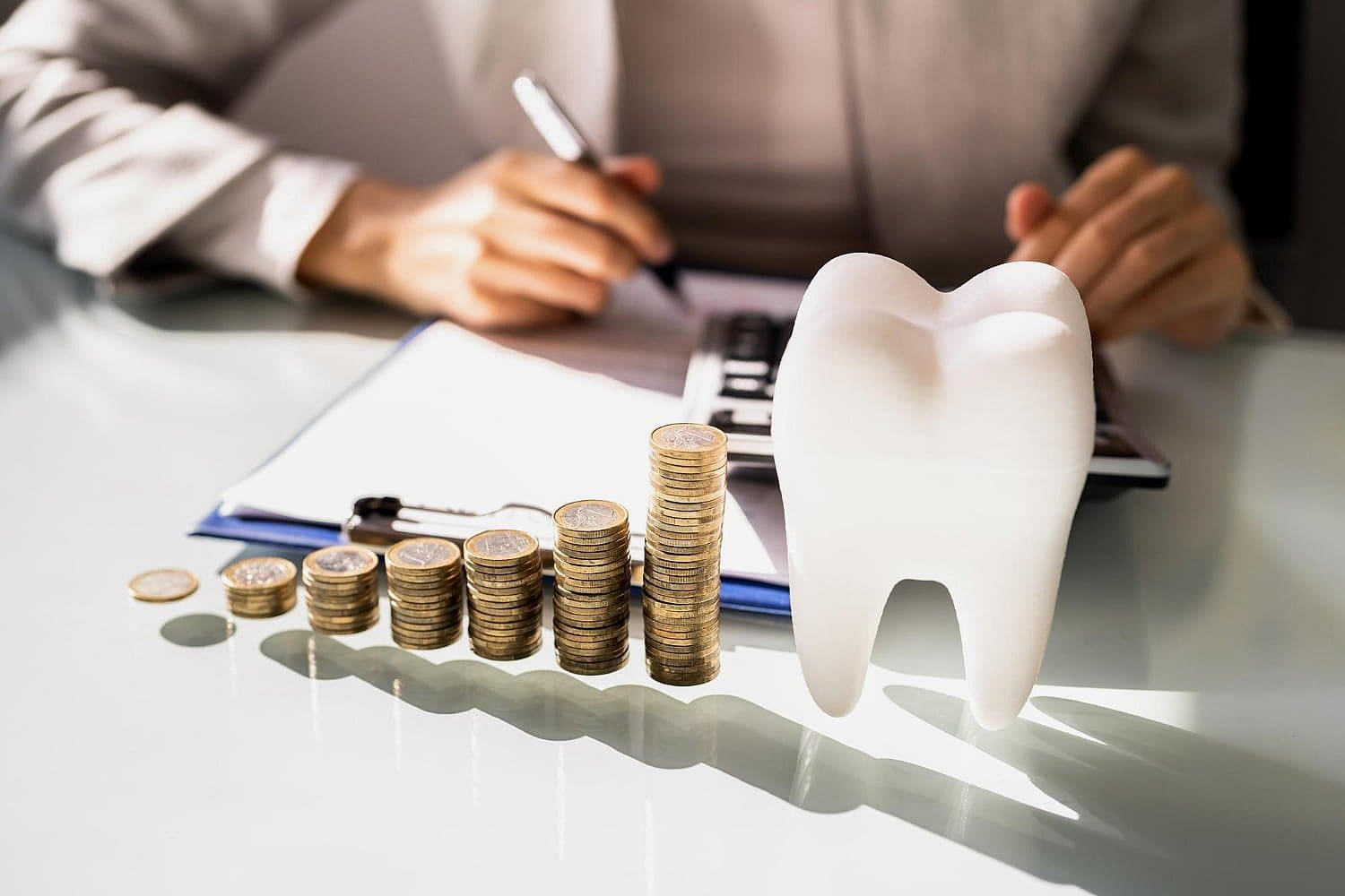 Stacked coins beside a large tooth model on a desk hint at affordability, with a person jotting down notes about affordable braces on a clipboard in the background in San Antonio, TX
