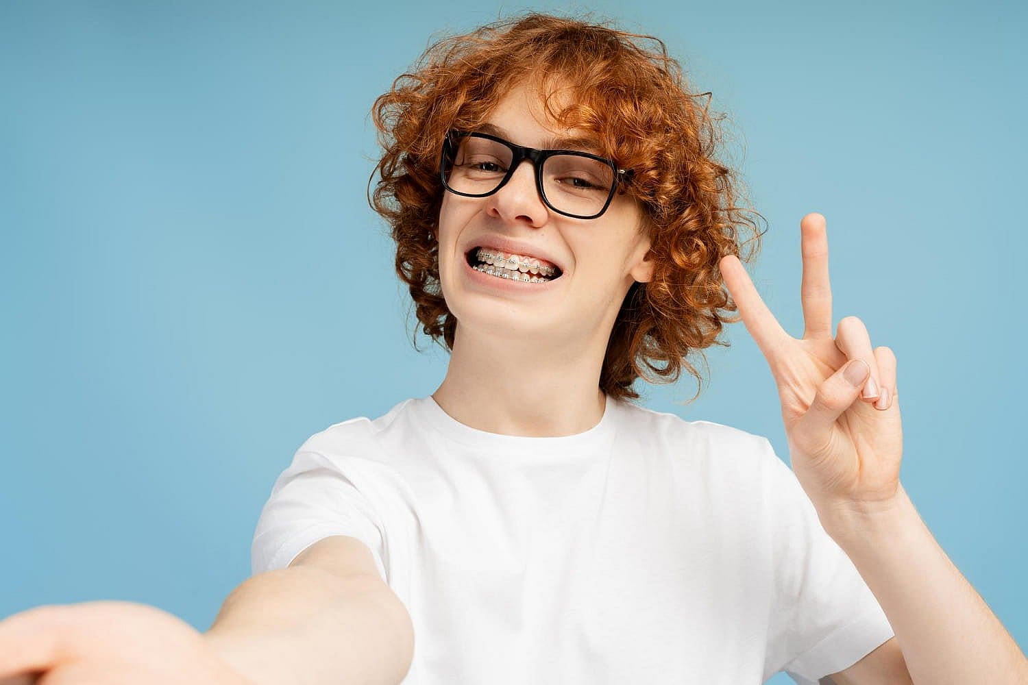 A smiling person with curly red hair, glasses, and affordable braces sports a white t-shirt, flashing a peace sign against a blue background in San Antonio, TX