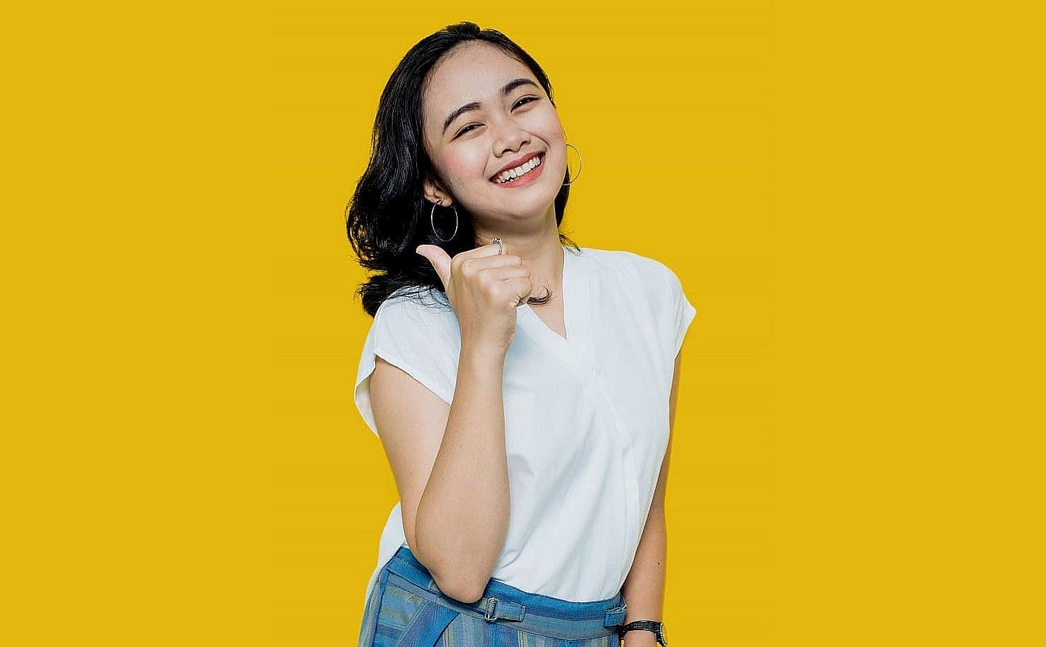 A woman in a white shirt and plaid skirt smiles, giving a thumbs-up against a bright yellow background, showcasing her perfect smile thanks to an Invisalign payment plan in San Antonio, TX