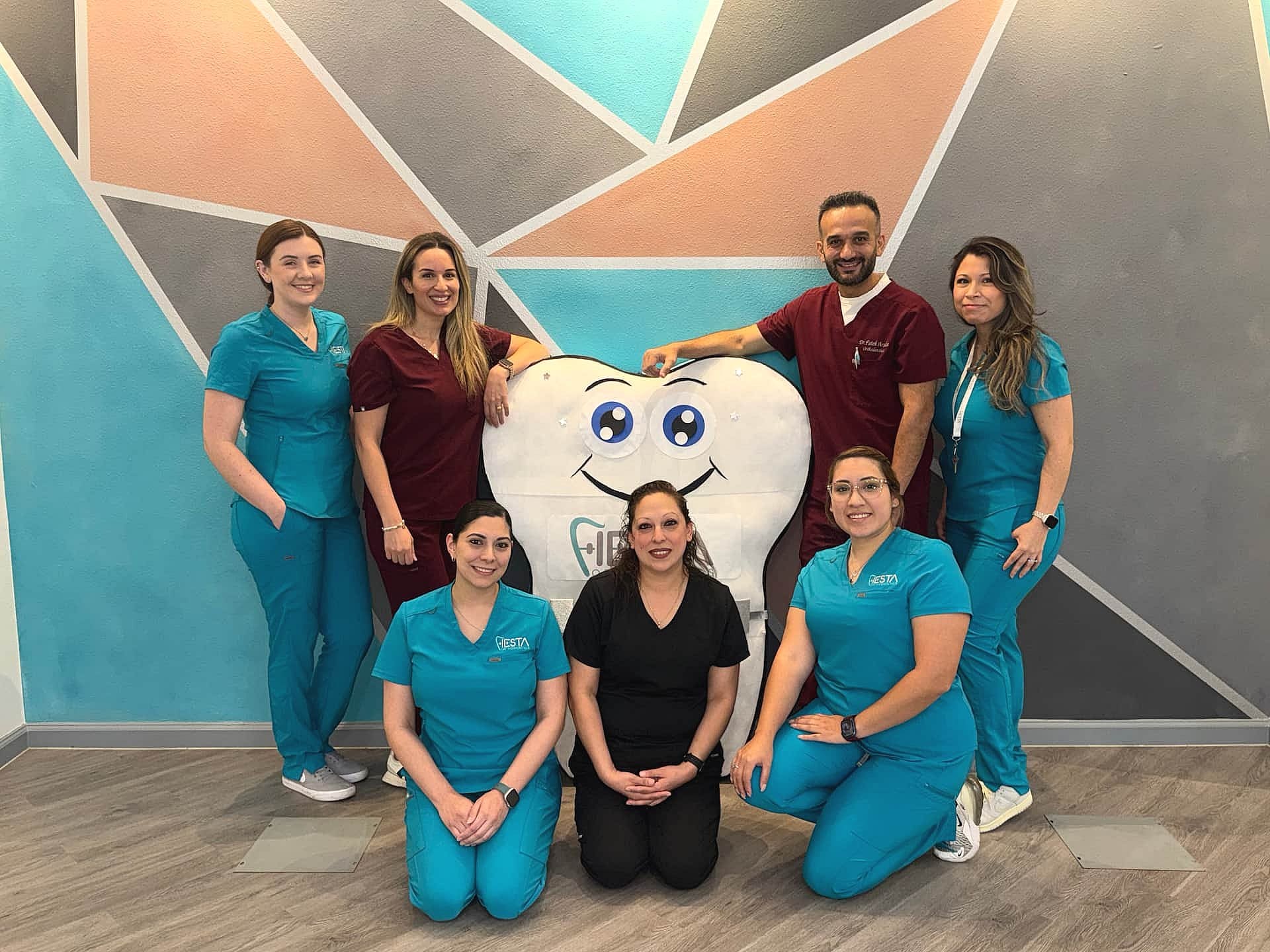 At Fiesta Orthodontics, six team members in scrubs and one in black clothing pose joyfully around a large cartoon tooth with a smiling face, all set against a striking geometric wall backdrop in San Antonio, TX after free orthodontic consultation