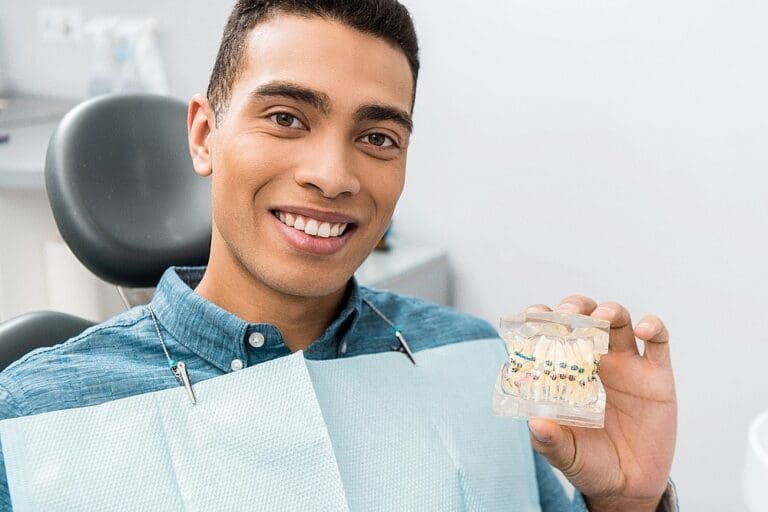 A person sitting in a dental chair smiles and holds a dental model with braces, wearing a blue shirt and dental bib, possibly wondering how long do braces take in San Antonio TX.