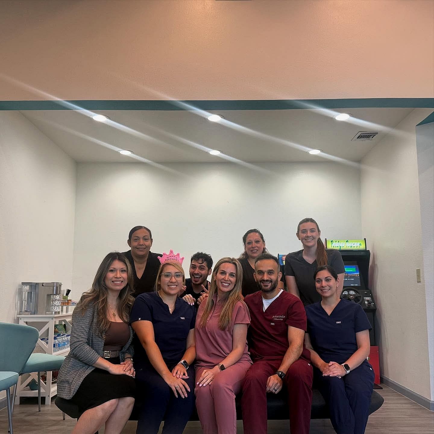 At Fiesta Orthodontics in West San Antonio, TX, nine people—some in medical scrubs and one with a pink crown—smile together in a brightly lit room featuring a coffee station and an arcade game.