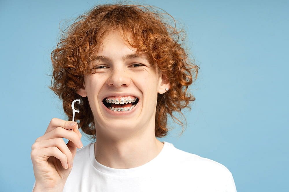 A person with curly hair and metal braces holds a dental floss pick in front of a blue background, representing Fiesta Orthodontics in East San Antonio, TX.