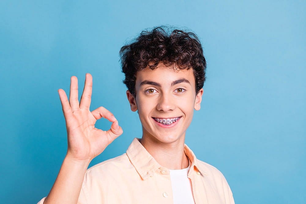 A smiling teenager with curly hair and metal braces flashes an "OK" hand gesture against a solid blue background at Fiesta Orthodontics in West San Antonio, TX.