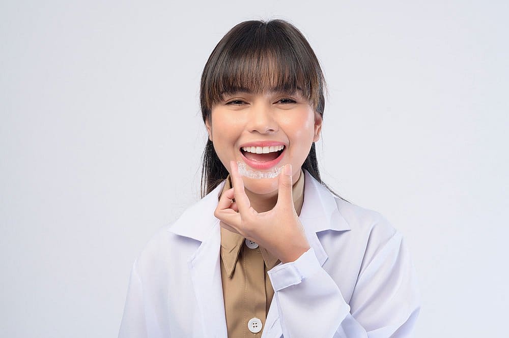 At Fiesta Orthodontics in East San Antonio, TX, a team member in a white lab coat smiles while displaying an Invisalign® Clear Aligner against a plain background.