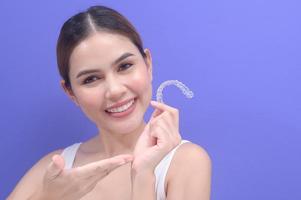 A woman smiles while holding Invisalign® Clear Aligners and gesturing toward them, set against a purple background at Fiesta Orthodontics in West San Antonio, TX.