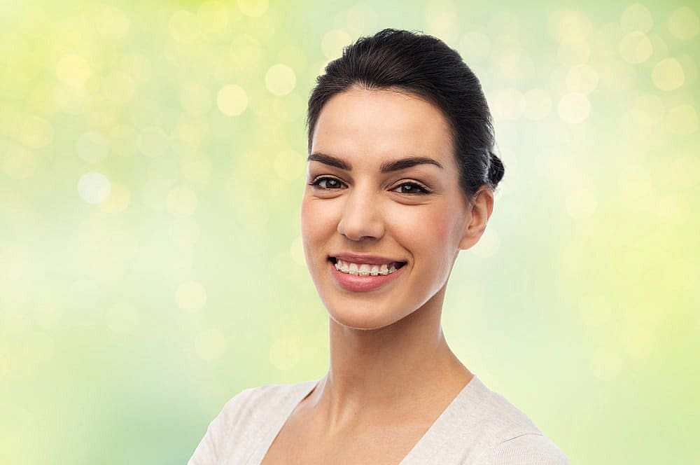A woman wearing a light-colored top smiles at the camera with clear braces, set against a blurred green and yellow background—capturing a welcoming moment at Fiesta Orthodontics in East San Antonio, TX.