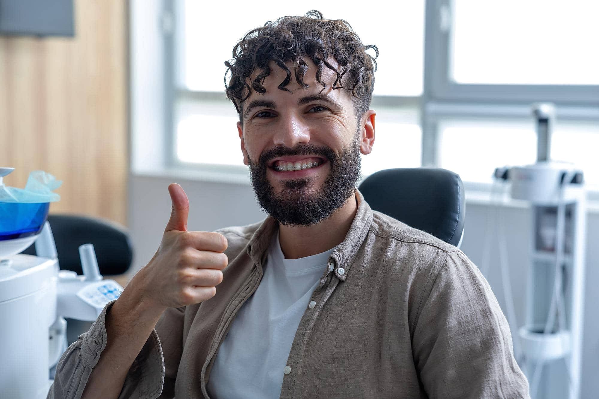 A man with curly hair and a beard, smiling and giving a thumbs up, sits in a dental office chair at Fiesta Orthodontics in West San Antonio, TX, with dental equipment and clear braces visible behind him.