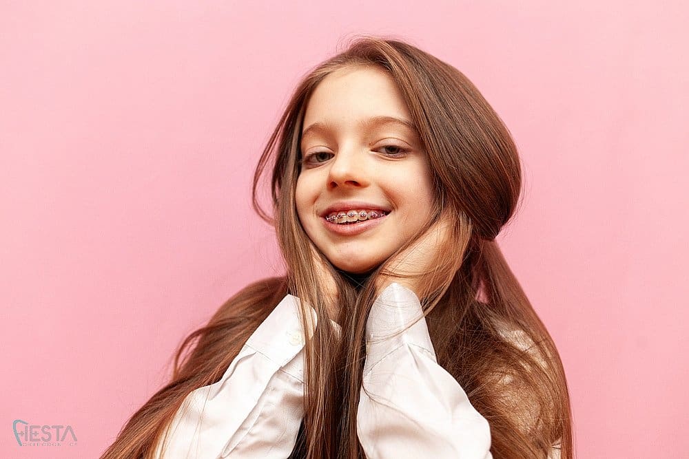 A girl with long brown hair and braces for overbite in San Antonio, TX while holding her face with both hands against a pink background at Fiesta Orthodontics.
