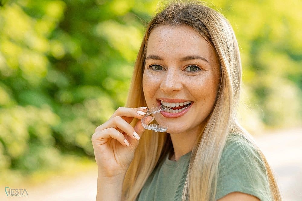 Young woman with long blonde hair smiling while holding a clear aligner outdoors, showcasing Invisalign treatment after understanding about Does Insurance Cover Invisalign in San Antonio, TX.