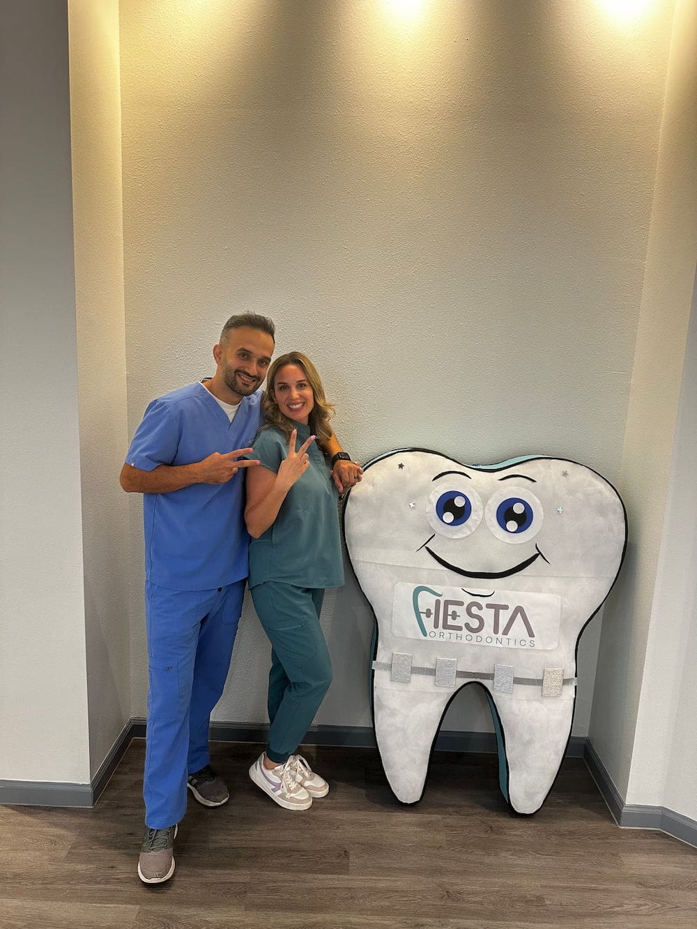 Smiling in scrubs, Dr. Fateh Arslan and Dr. Ayla Idilbi pose with peace signs by a Fiesta Orthodontics sign in San Antonio, TX.