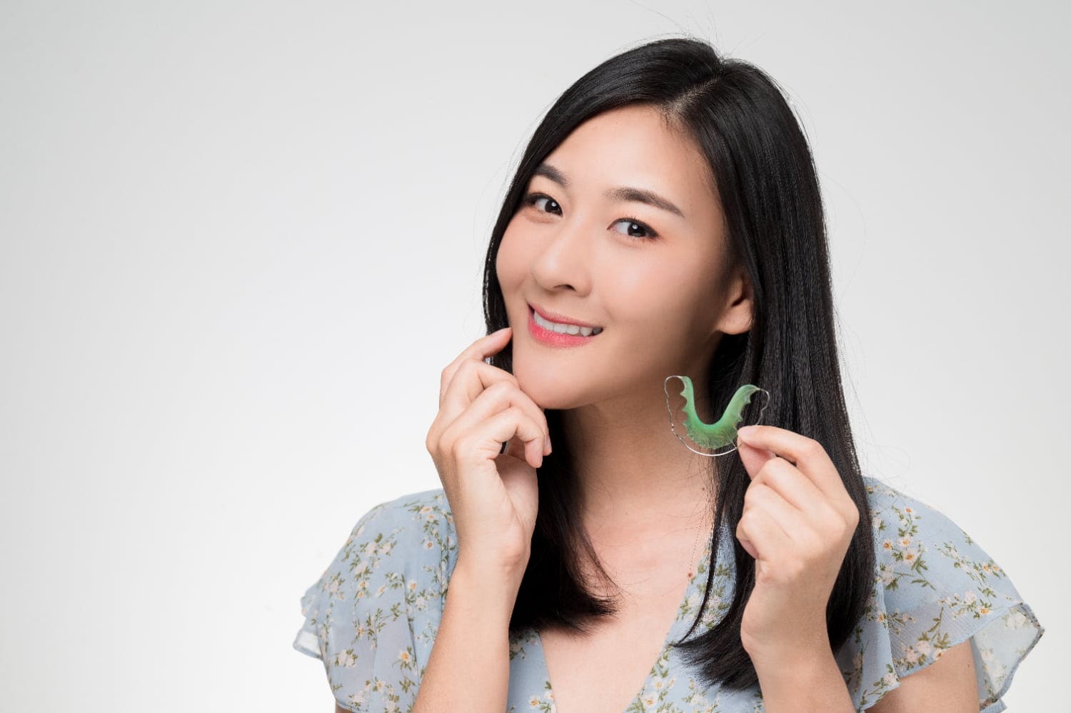Smiling woman in a floral top holds a green dental retainer of orthodontic appliances at Fiesta Orthodontics in West San Antonio and East San Antonio, TX.