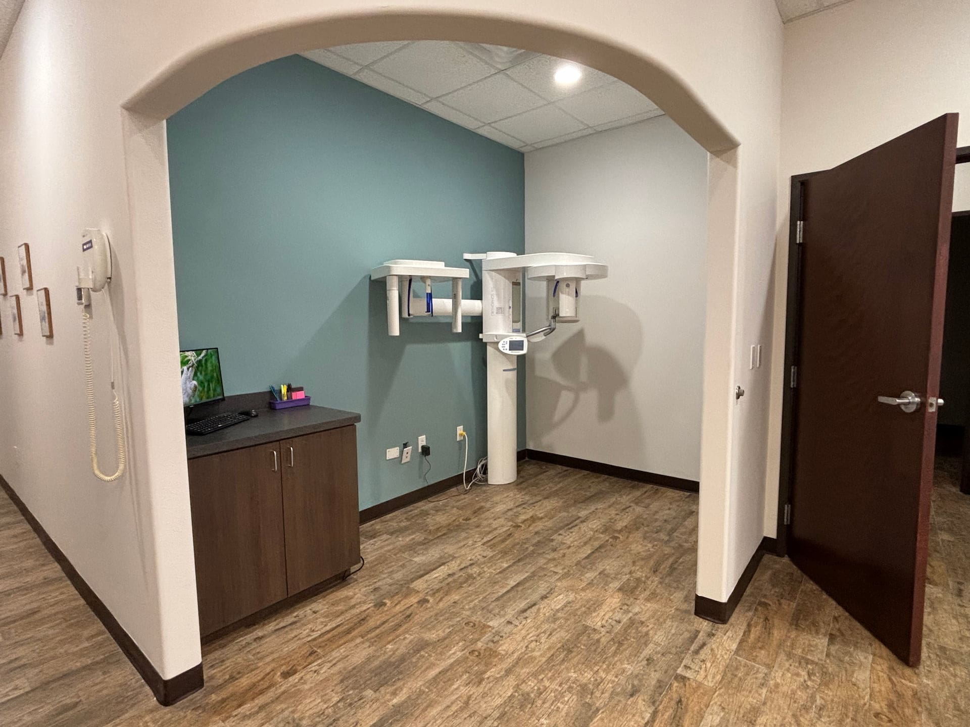 At Fiesta Orthodontics in East San Antonio, TX, a panoramic X-ray machine stands by a brown cabinet and open wood door in the exam room.