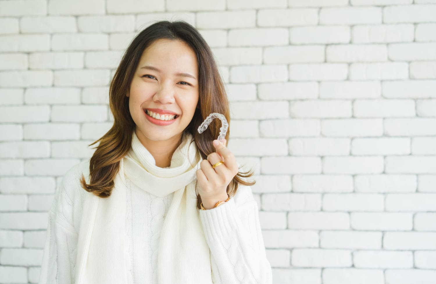 A woman in a white sweater smiles, holding an Invisalign aligner at Fiesta Orthodontics in West San Antonio and East San Antonio, TX.