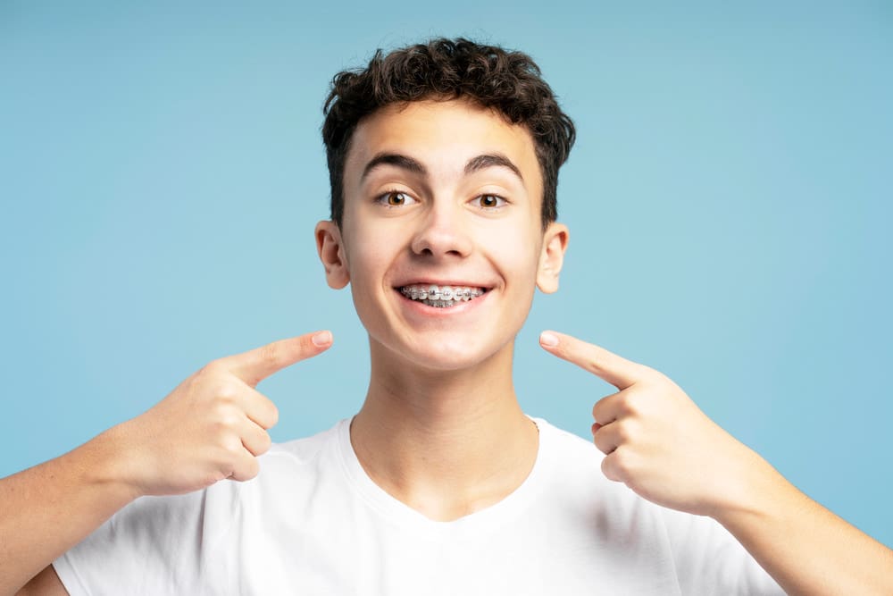Smiling teen boy with dark curly hair points to new braces, wearing a white t-shirt; Fiesta Orthodontics in East San Antonio, TX.