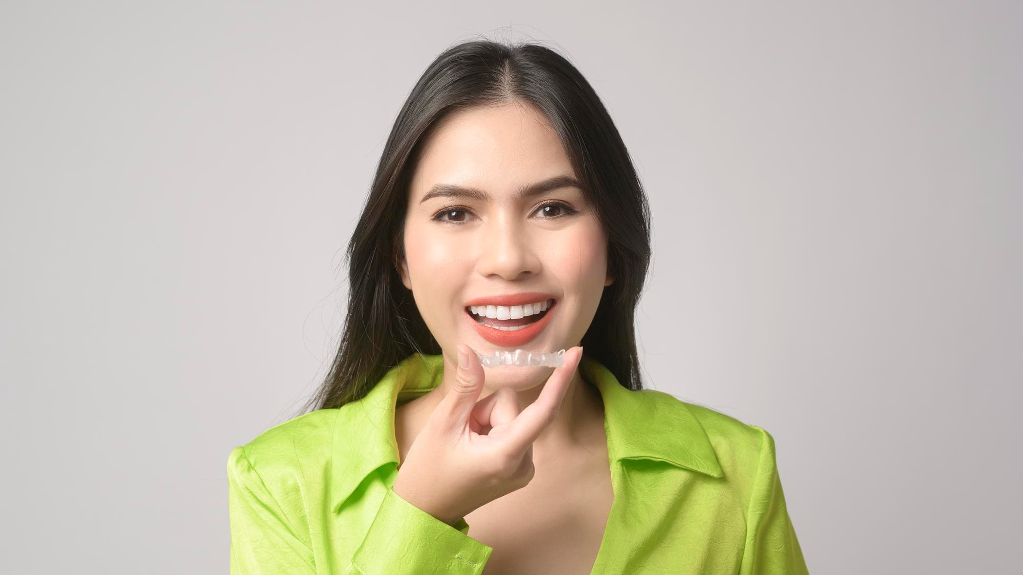 Smiling woman in bright green shirt holds a Invisalign® Clear Aligners by her mouth at Fiesta Orthodontics in East San Antonio, TX.