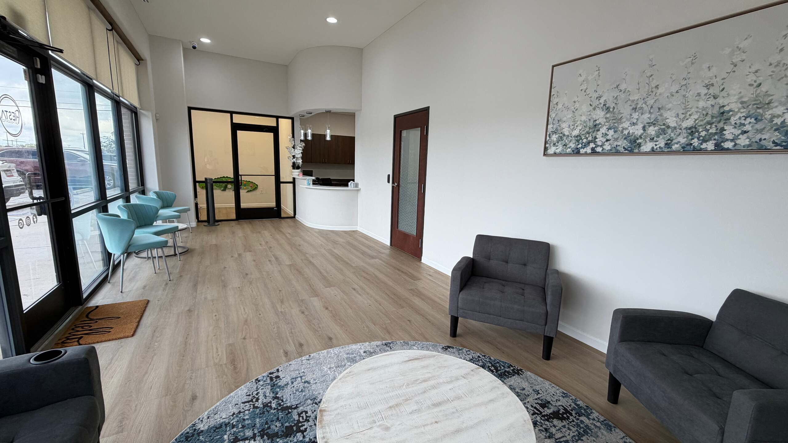 Waiting room with teal chairs by the front windows, gray armchairs, a round coffee table and the reception desk at Fiesta Orthodontics in East San Antonio, TX.
